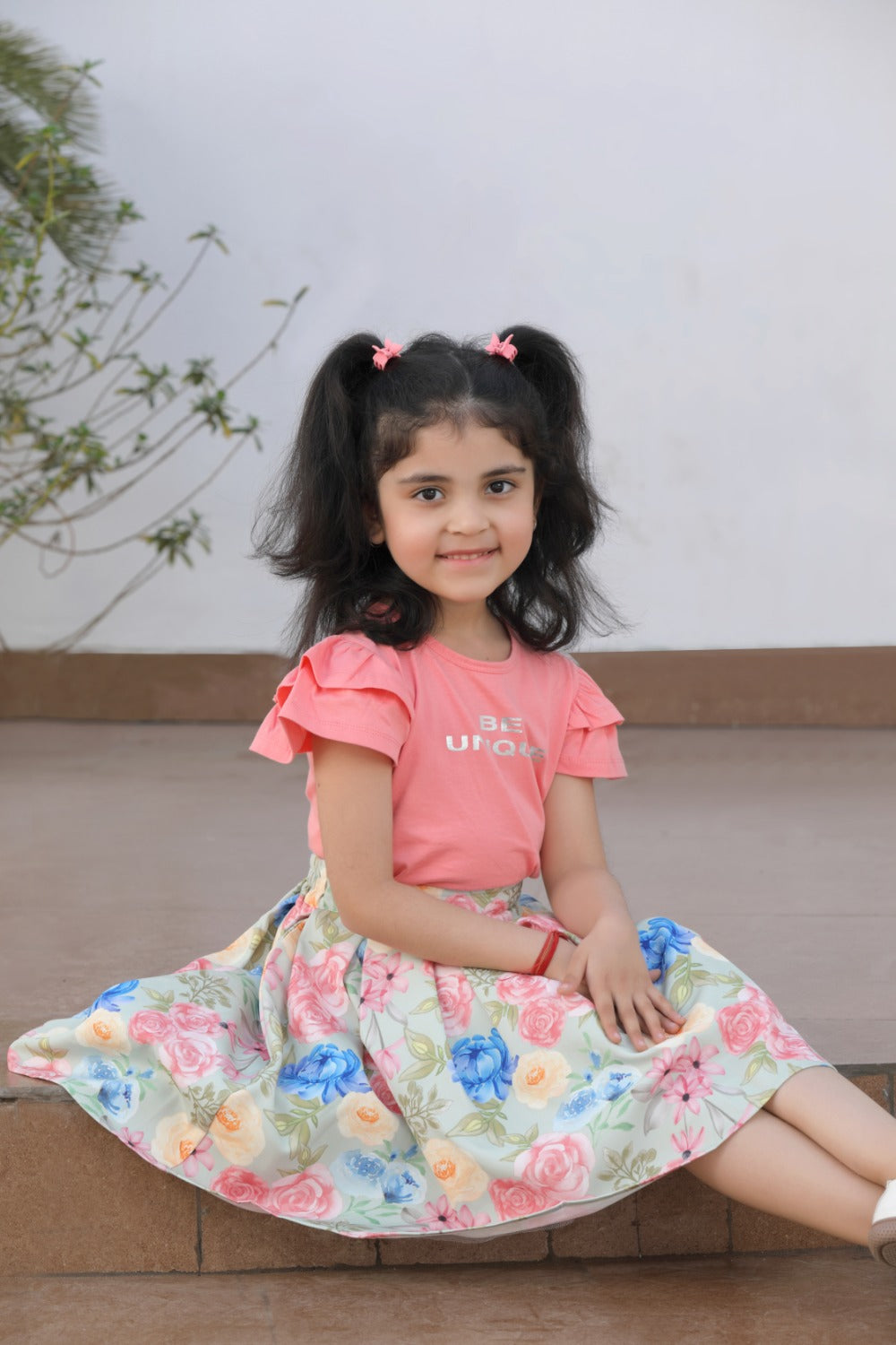 Girl in coral ruffle top and floral skirt seated, smiling
