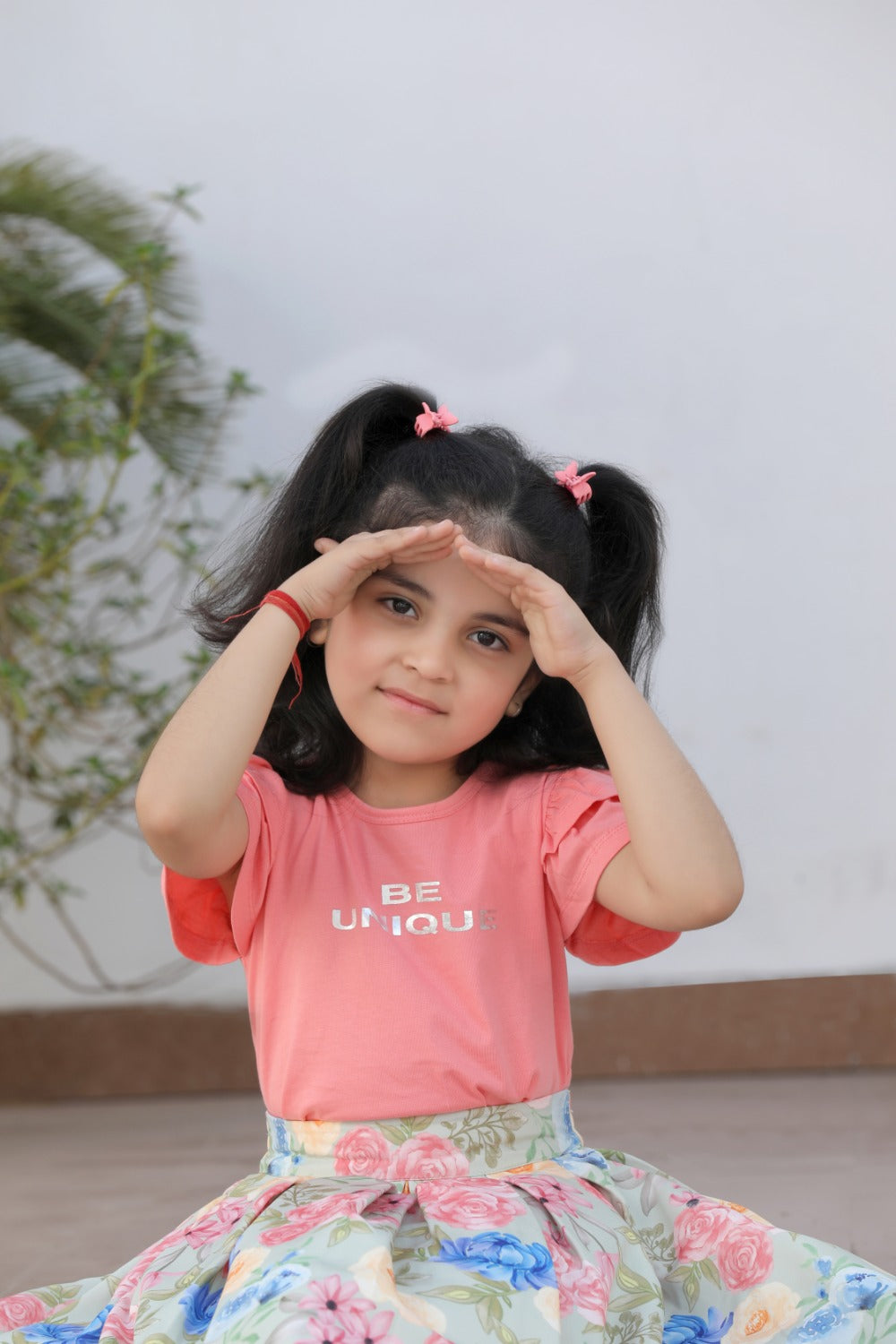 Girl in coral tee and floral skirt posing outdoors, hands on forehead